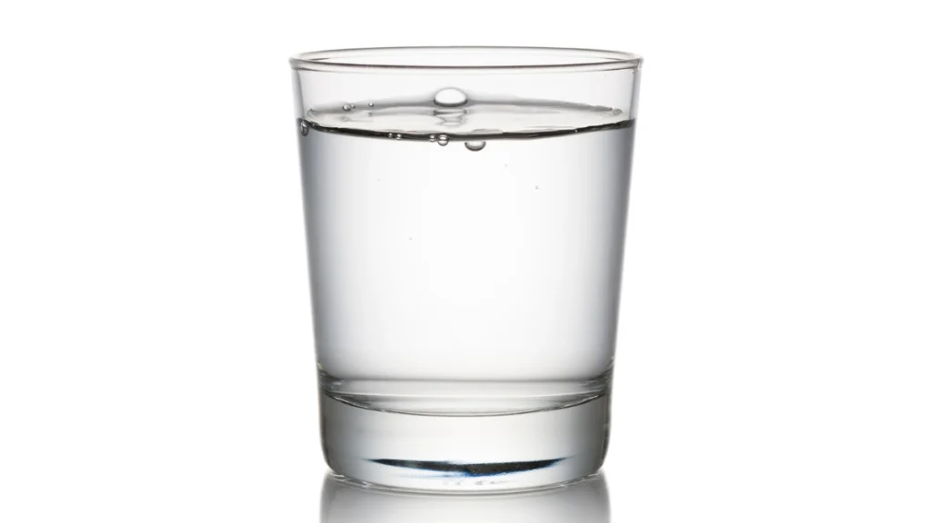 A clear glass of water on a table, with a hand holding a bottle in the background
